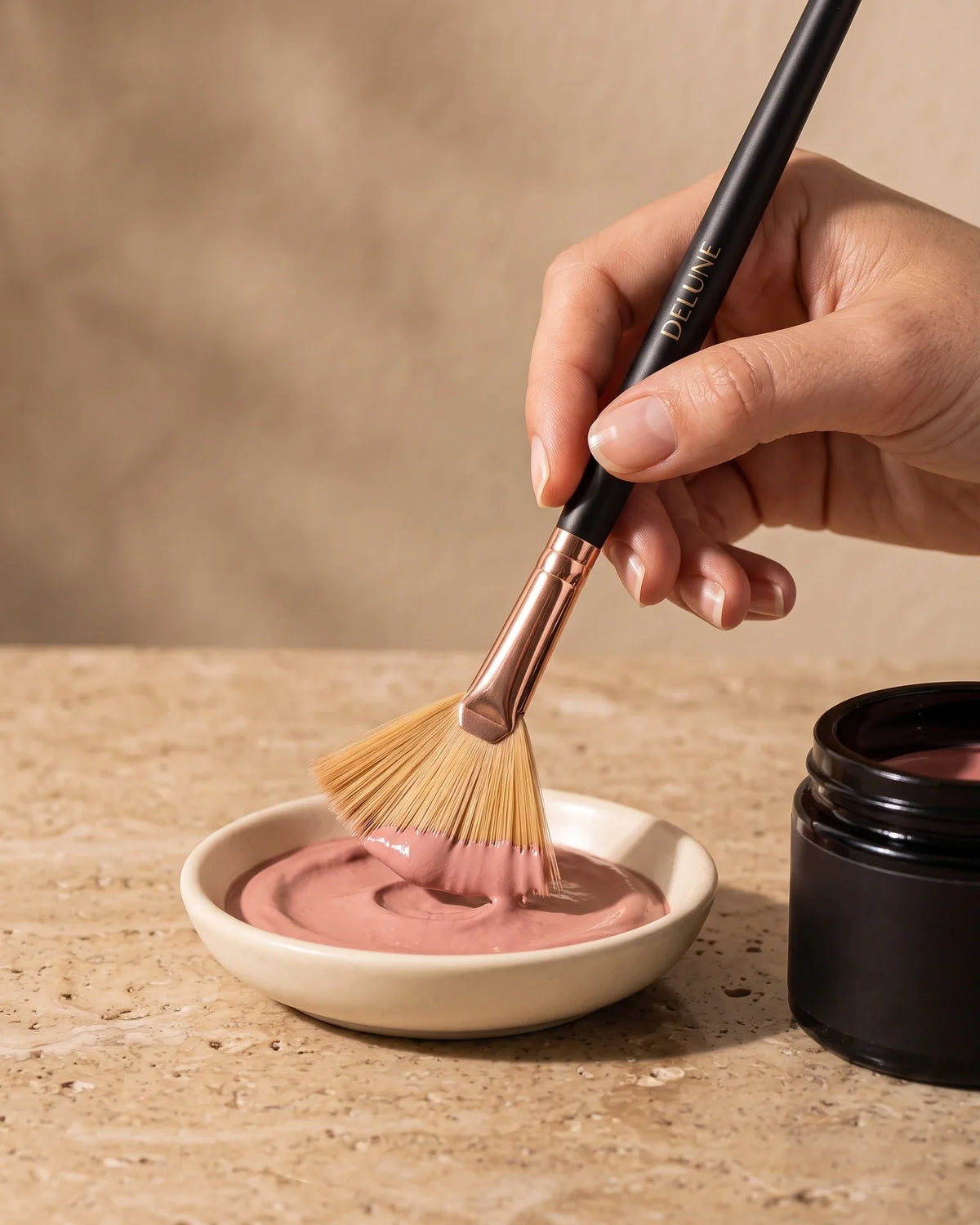 Hånd, der tager rosa lerpasta på Delunes viftepensel fra en keramikskål