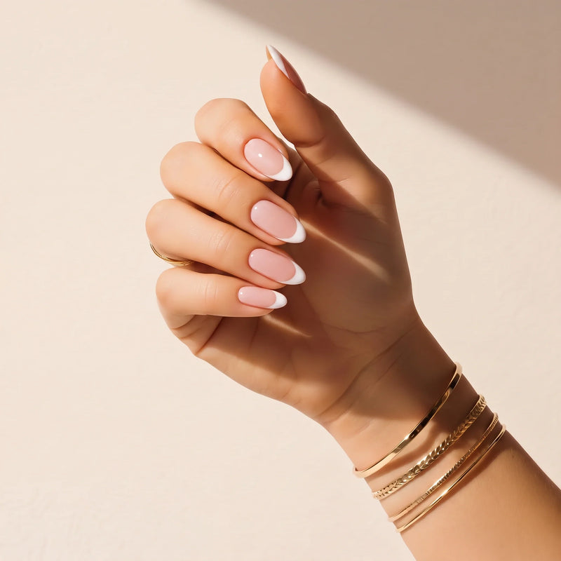 Hånd med Delune French Manicure – delvist transparente neglefolier uden UV og guldarmbånd mod en varm pudset væg