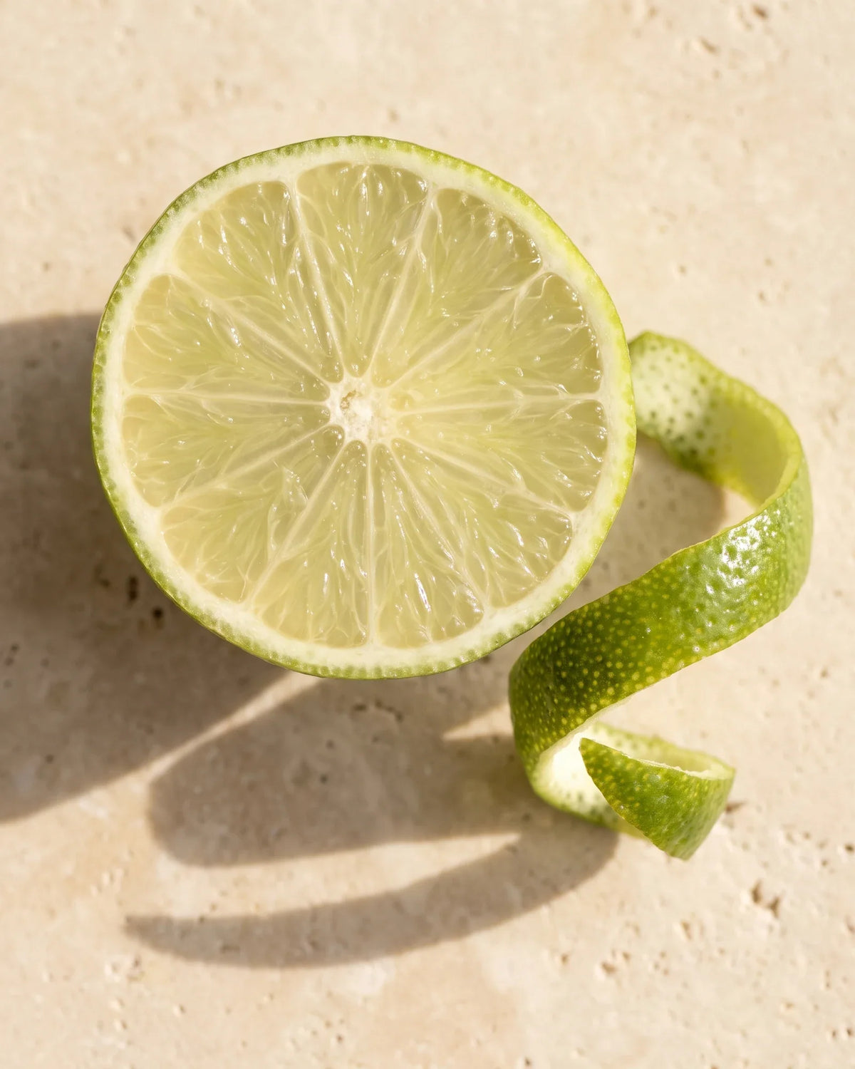 En halveret frisk lime og en krølle limeskal set ovenfra på varm travertin – den skarpe, friske duft fra Delune Lime – ren æterisk olie