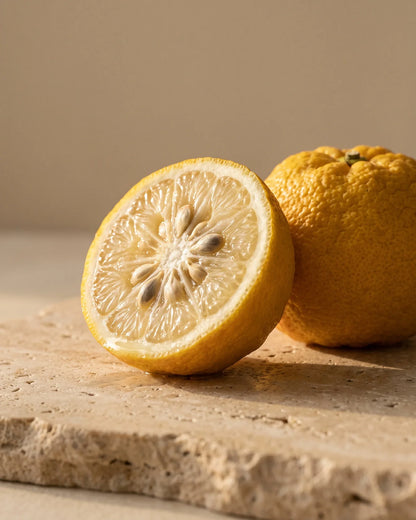 En halveret yuzu ved siden af en hel, knudret yuzu på varm travertin – den klare, blomstrede citrusduft i Delunes rene æteriske olie af yuzu