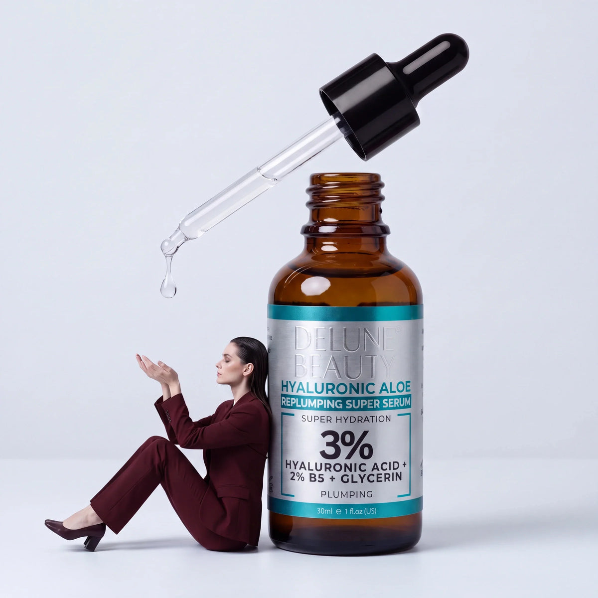 Pleje: en kvinde i oxblodsrødt jakkesæt læner sig op ad et kæmpe Delune Hyaluronic Aloe-serum og griber en faldende dråbe
